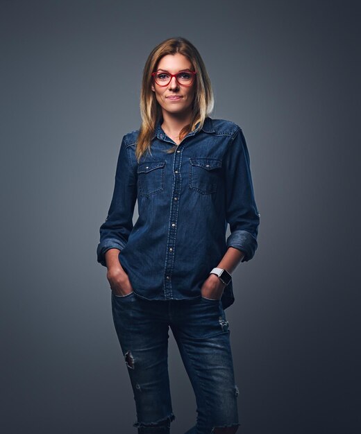 Blond female dressed in denim shirt and red eyeglasses posing over grey background.