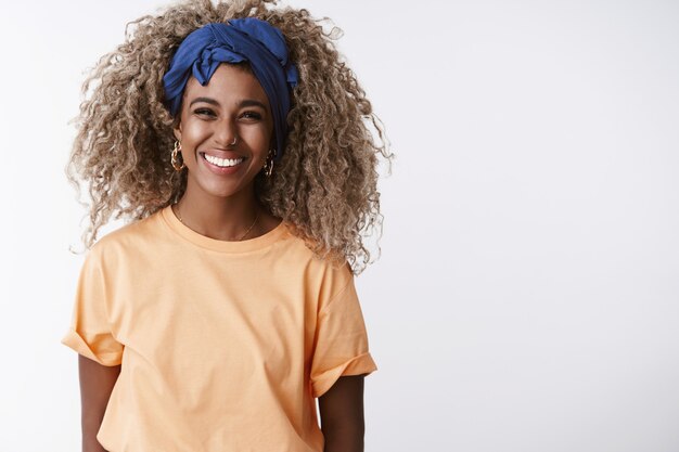 Blond girl with afro hairstyle, stylish headband and orange t-shirt, laughing and smiling joyful, have fun standing white wall