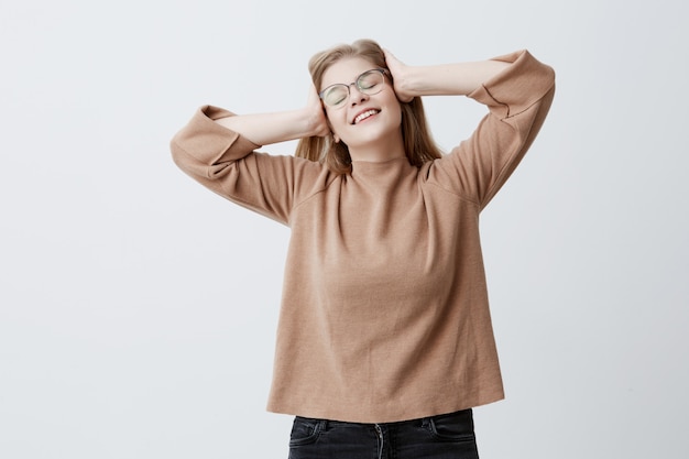 Blond joyful caucasian girl wearing brown sweater and eyeglasses, holding hands on head while having fun indoors against grey studio wall background, smiling broadly, demonstrating white even teeth