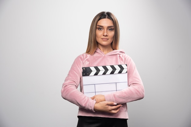 Blond lady in pink sweatshier holding a blank clapper board and gives natural poses.