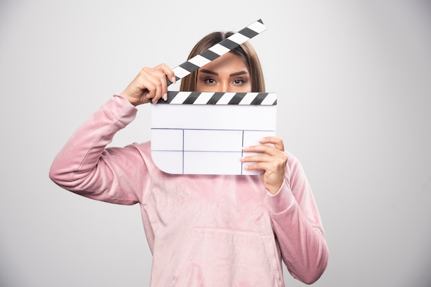 Free photo blond lady in pink sweatshier holding a blank clapper board and looking through it.