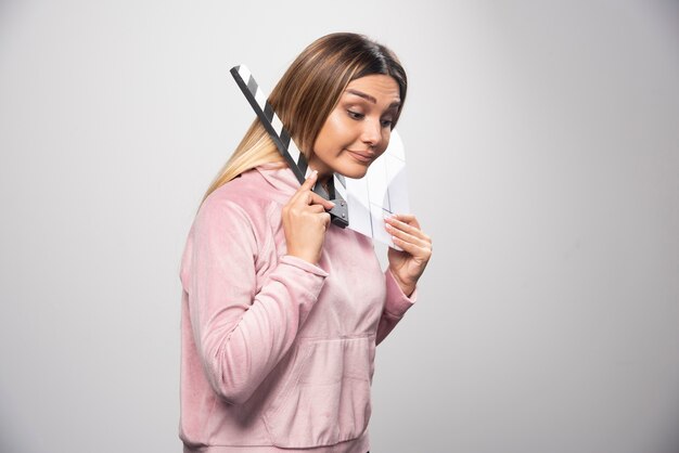 Blond lady in pink sweatshier holding a blank clapper board and puts a part of her body through it.