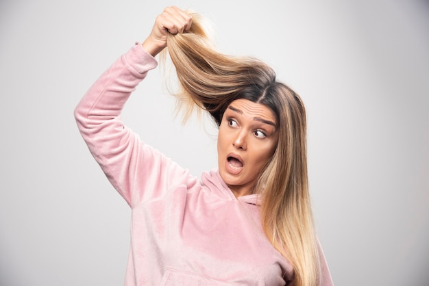 Blond lady in pink sweatshirt feels dissatisfied about her dry hair or hair color.