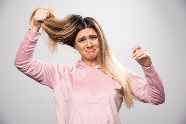 Blond lady in pink sweatshirt feels dissatisfied about her dry hair or hair color.