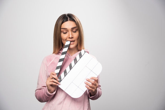 Free photo blond lady in pink sweatshirt holding a blank clapper board and bites a corner of it