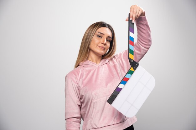 Blond lady in pink sweatshirt holding a blank clapper board and gives positive and fun poses.