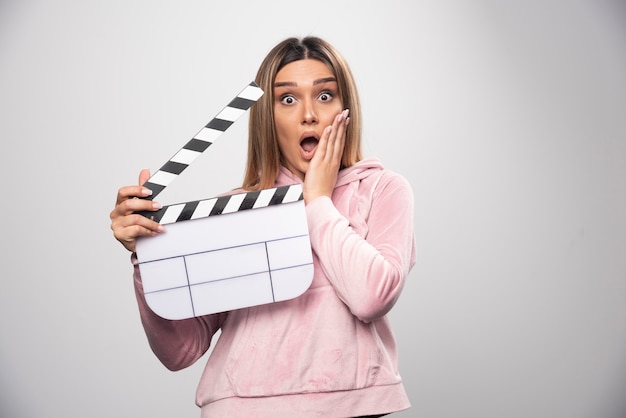 Free photo blond lady in pink sweatshirt holding a blank clapper board looks surprized and confused.