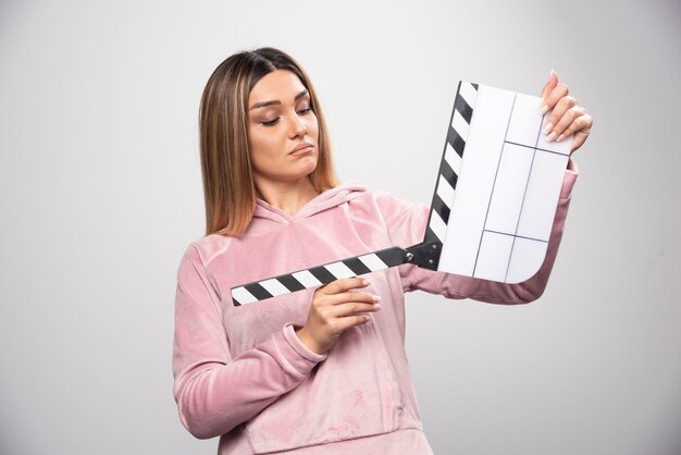 Blond lady in pink sweatshirt holding a blank clapper board and trying to understand what is it.