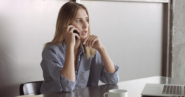 Blond model in a relaxed buttonup shirt switches between her laptop and her phone at work