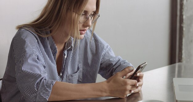 Блондинка-модель в расслабленной рубашке на пуговицах переключается между ноутбуком и телефоном на работе.