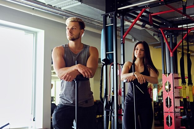 Blond muscular male and athletic brunette female hold a barbell in a gym club.