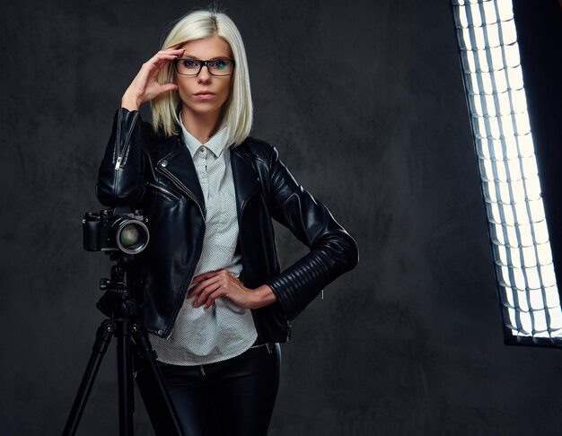 A blond photographer female dressed in a black leather jacket holds professional digital camera and a tripod.