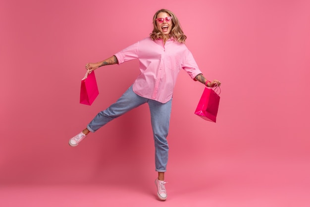 ピンクのシャツとジーンズの金髪のきれいな女性がショッピングバッグを持って楽しんで分離されたピンクの背景にジャンプして笑っている