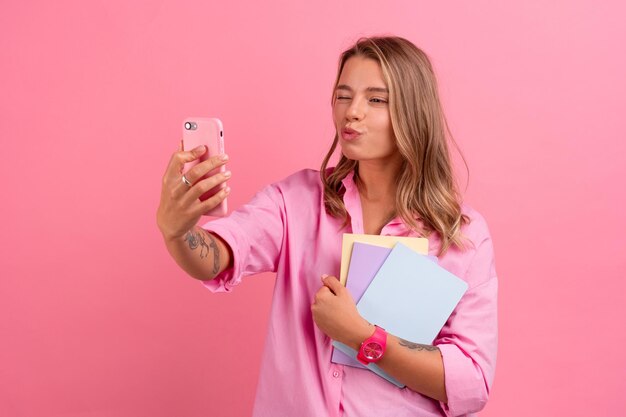 Blond pretty woman in pink shirt smiling holding holding notebooks and using smartphone