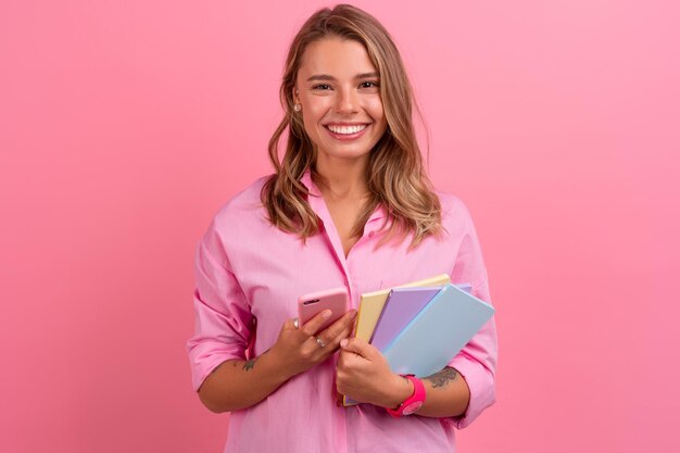 Blond pretty woman in pink shirt smiling holding holding notebooks and using smartphone