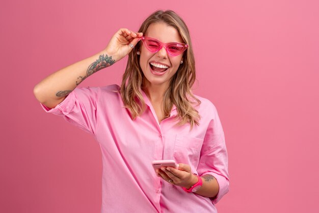 핑크색 셔츠를 입은 금발의 예쁜 여성이 선글라스를 끼고 즐겁게 웃고 있는 분홍색 외진 곳에 스마트폰을 들고 웃고 있다