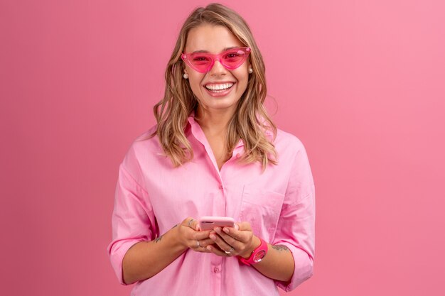 Bella donna bionda in camicia rosa sorridente che tiene tenendo lo smartphone in posa su rosa isolato sorridente divertendosi indossando occhiali da sole