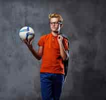 Free photo a blond teenager boy dressed in a jeans and orange t shirt holds a soccer ball over grey background.