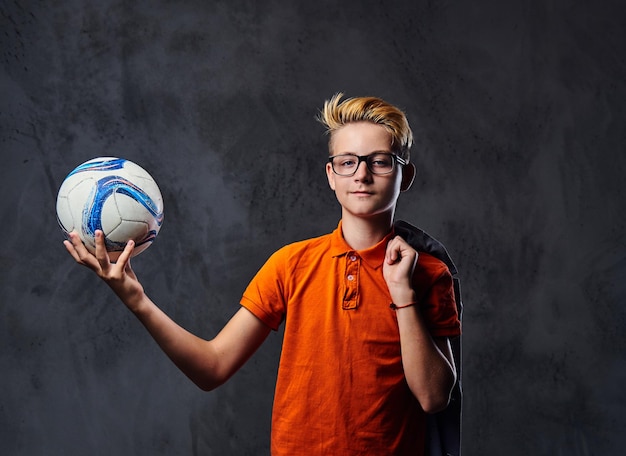 A blond teenager boy dressed in a jeans and orange t shirt holds a soccer ball over grey background.