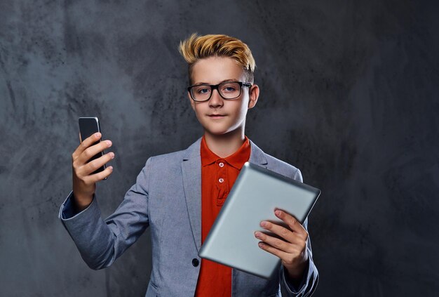 Белокурый подросток в очках держит планшетный ПК и смартфон.