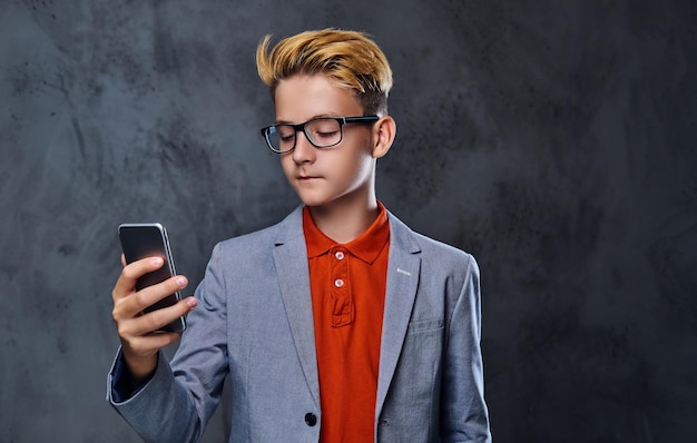 Il ragazzo biondo dell'adolescente in una giacca e occhiali da vista tiene lo smartphone.