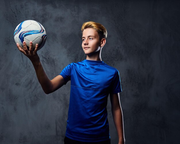 Blond teenager, soccer player dressed in a blue uniform holds a ball.