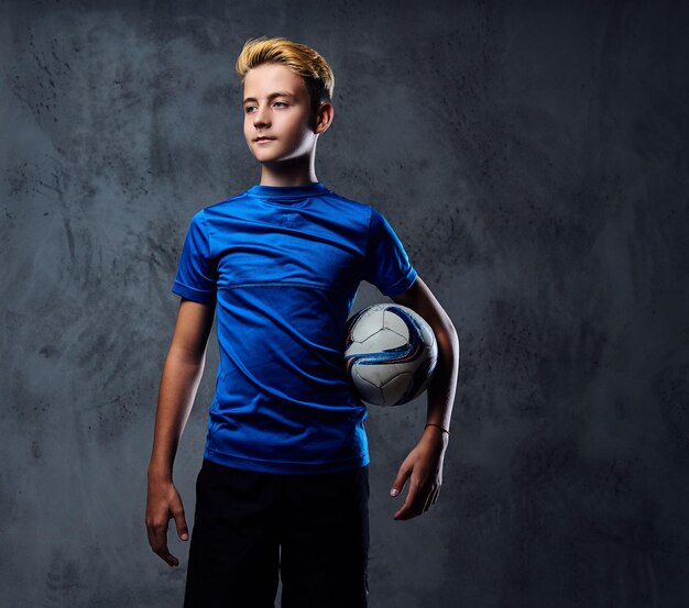 Blond teenager, soccer player dressed in a blue uniform holds a ball.