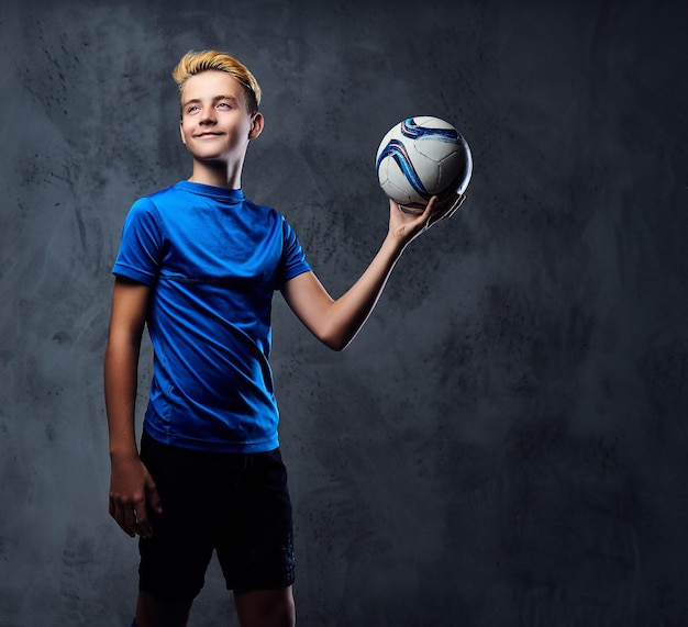 Blond teenager, soccer player dressed in a blue uniform holds a ball.