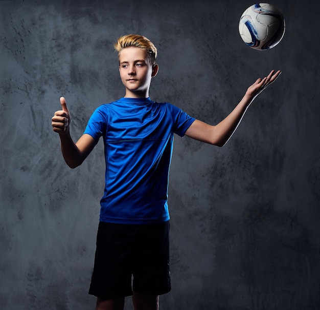 Blond teenager, soccer player dressed in a blue uniform plays with a ball.