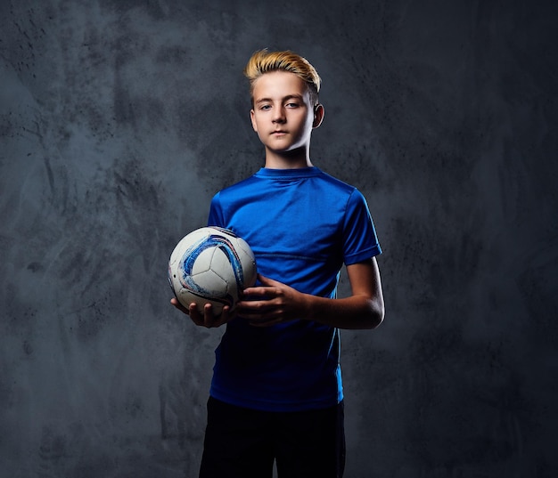 Blond teenager, soccer player dressed in a blue uniform plays with a ball.