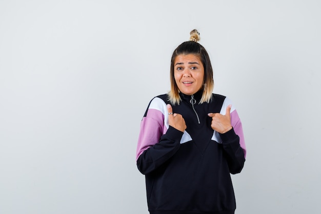 Blond woman pointing at herself with fingers in black tracksuit and looking surprised