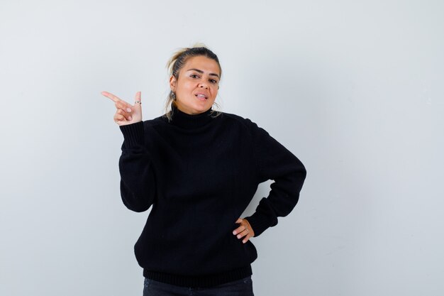 Blond woman pointing up, with hand on waist in black sweater and looking displeased , front view.