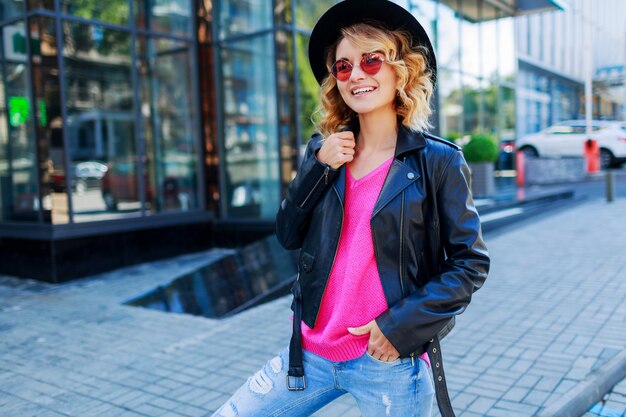 blond woman posing on modern streets. Stylish autumn outfit, leather jacket and knitted sweater. Pink sunglasses.
