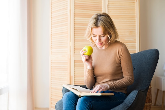 Blond woman reading book while eating apple