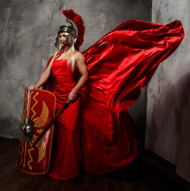 Blond woman in Roman red fluttering dress holds sword and shield.