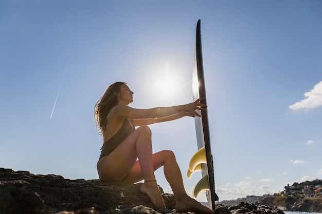 Foto gratuita donna bionda in costume da bagno che si siede sulla riva del mare roccioso con tavola da surf