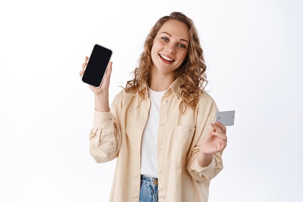 巻き毛のブロンドの女性、空のスマートフォンの画面とクレジットカードを表示、インターネットで購入、アプリのデモ、白い壁