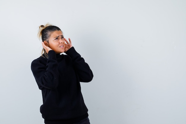 Blond woman with hands behind ears, standing sideways in black sweater and looking gloomy .