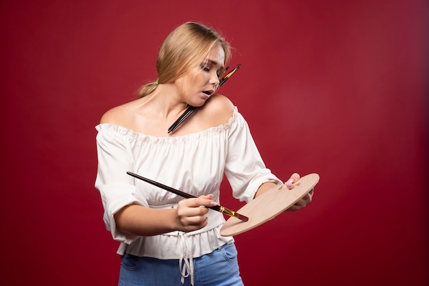 Free photo blonde artist holds a palette and brushes and looks positive and dedicated.