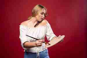 Free photo blonde artist holds a palette and brushes and looks positive and dedicated.