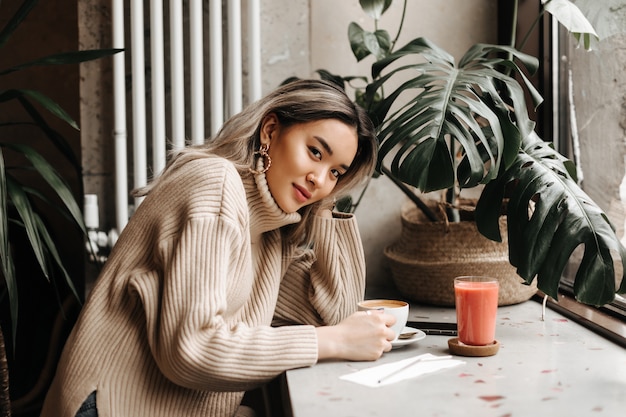 Free photo blonde asian woman in beige oversized sweater sits in cafe with cup of coffee and carrot juice