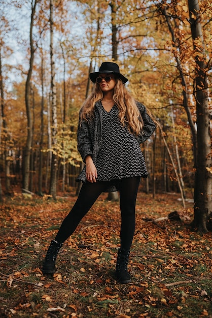 Foto gratuita modello femminile attraente biondo con cappotto nero e cappello in posa in una foresta autunnale