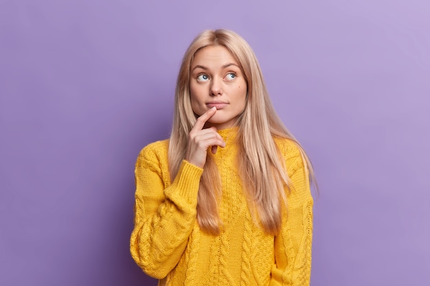 Blonde attractive young European woman keeps finger on lips looks with pensive expression above makes important decision builds plans in mind wears yellow sweater 