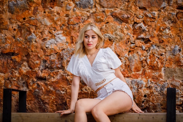 Blonde beautiful girl is looking away by sitting on bench on stone background