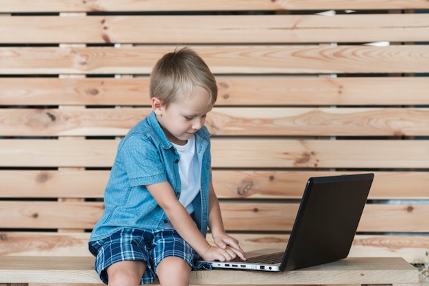 Foto gratuita ragazzo biondo che si siede sulla tabella facendo uso del computer portatile