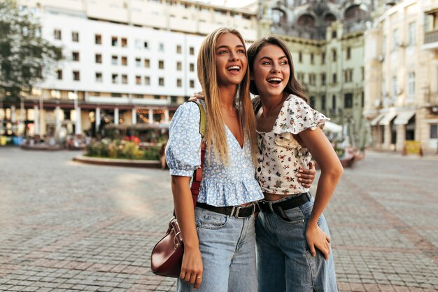 Blonde and brunette women in stylish outfits look away in good mood