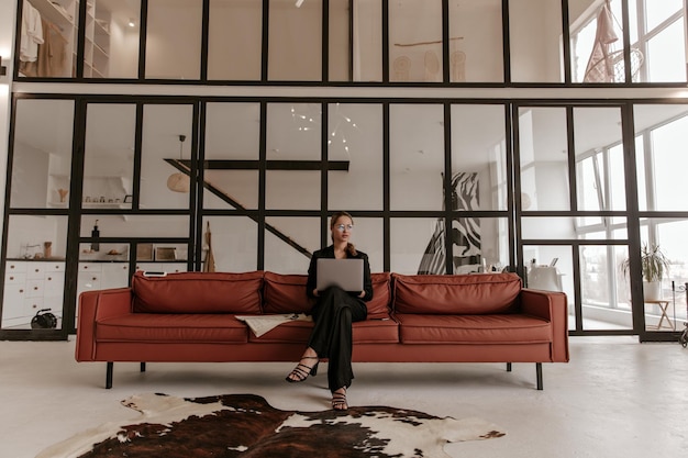 Free photo blonde charming woman in silk black jumpsuit and stylish eyeglasses sits on brown big soft sofa ho