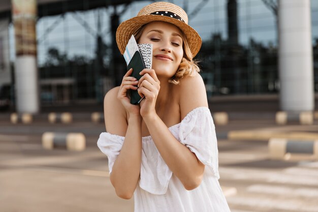 Blonde cheerful woman in boater and white dress holds passport and smiles sincerely