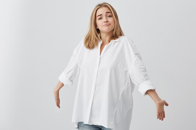 Blonde confused caucasian female in white shirt shrugging shoulders, stretching arms forward, staring  with questionable and clueless look, saying I don't know. People and emotions concept.