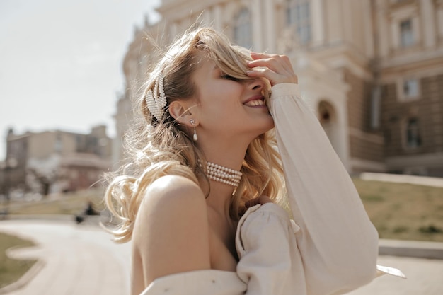 Blonde curly young woman smiles sincerely outdoors Happy charming girl in white blouse and pearl necklace poses in city center